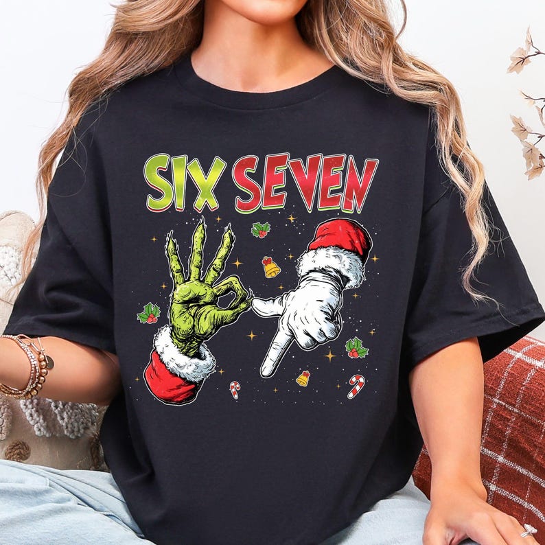 Puede incluir: Camiseta negra con un gr&aacute;fico de dos manos, una verde y otra blanca, formando los n&uacute;meros seis y siete. El texto "SIX SEVEN" est&aacute; en verde y rojo. Los gr&aacute;ficos festivos de acebo, campanas y bastones de caramelo completan el dise&ntilde;o.