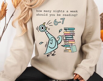 Puede incluir: Sudadera beige con una ilustración de paloma de dibujos animados y el texto "how many nights a week should you be reading?" con los números "6-7". La paloma es azul claro con detalles en negro, y una pila de libros está a la derecha.