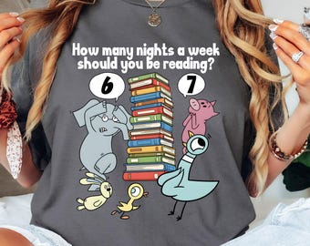 Puede incluir: Camiseta gris con el texto "How many nights a week should you be reading?" e ilustraciones de animales. Una pila de libros está en el centro.