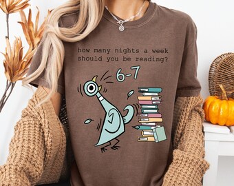 Puede incluir: Camiseta marrón con una ilustración de paloma de dibujos animados y texto. La paloma es azul claro, y el texto dice "how many nights a week should you be reading?" y "6-7". También se muestra una pila de libros.
