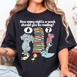 May include: Black t-shirt with the text "How many nights a week should you be reading?" and illustrations of an elephant, pig, pigeon, and other animals. The design includes a stack of books and the numbers 6 and 7.