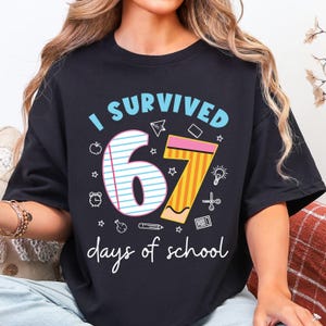 May include: Black t-shirt with the text "I SURVIVED 67 days of school." The number 67 is designed with a notebook and pencil. The shirt has a crew neck and short sleeves.
