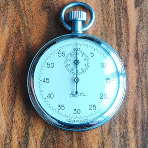 Op de afbeelding: Een vintage stopwatch met een zilverkleurige metalen behuizing en een witte wijzerplaat. De wijzerplaat heeft zwarte cijfers, een kleinere binnenwijzerplaat en het woord "Azam". De tekst "СДЕЛАНОВ СССР" staat onderaan.