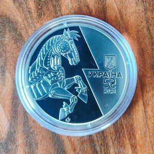 May include: A silver coin featuring a detailed mechanical horse design. The coin has the text "УКРАЇНА" and "5" along with the year "2024". The coin is encased in a clear protective case.
