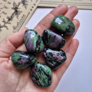 May include: Five polished tumbled stones featuring a mix of vibrant green zoisite, deep black hornblende, and patches of magenta ruby. Each stone has a smooth, glossy surface with irregular colour patterns and rounded edges, displayed together.