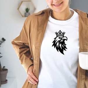 以下が含まれることがあります： 白いTシャツに、黒と白のグラフィックで描かれた、王冠をかぶった鷲のイラスト。鷲は右を向いており、翼を広げています。