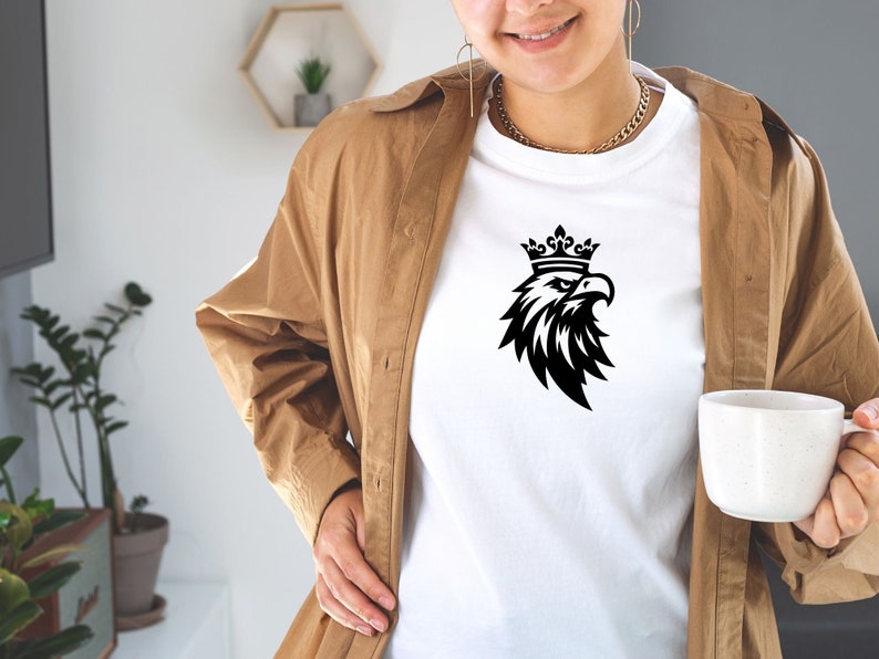 以下が含まれることがあります： 白いTシャツに、黒と白のグラフィックで描かれた、王冠をかぶった鷲のイラスト。鷲は右を向いており、翼を広げています。