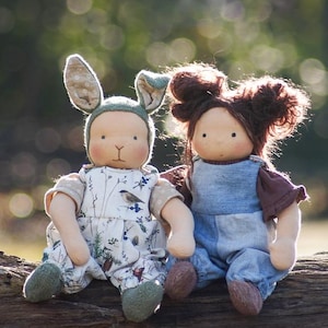 Peut inclure: Deux poupées en tissu faites à la main assises sur une bûche. La poupée de gauche porte un chapeau de lapin vert et une robe à fleurs blanche et verte. La poupée de droite porte une robe en jean bleu avec une chemise marron.