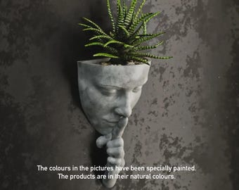 May include: A gray concrete wall planter shaped like a human face, with a finger pressed to the lips in a 'shush' gesture. The planter holds a green succulent plant. The text at the bottom says, "The colours in the pictures have been specially painted. The products are in their natural colours."