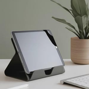 Puede incluir: Soporte negro para tableta con una tableta con pantalla en blanco. Un lápiz óptico blanco y un teclado están sobre la superficie blanca. Una planta en maceta está en el fondo.