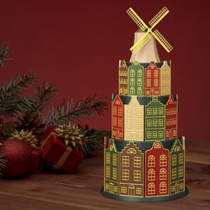 Peut inclure: Un moulin à vent décoratif à trois niveaux avec une série de maisons colorées. Les maisons sont peintes dans des tons de vert, de rouge et de beige, avec des accents dorés. Les pales du moulin sont dorées. La scène se déroule sur une surface en bois avec des décorations de Noël.