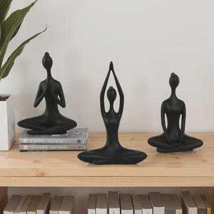 May include: Three black yoga pose figurines on a wooden table. The figurines are in different poses, with one in a seated position with arms raised. A potted plant and books are also on the table.