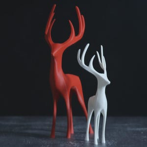 May include: Two deer ornaments, one red and one white, with elegant, slender forms and prominent antlers. They stand on a reflective surface against a dark backdrop. The design is minimalist and contemporary, suitable for home decor.