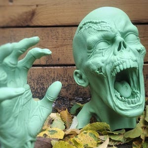 May include: A green zombie head and hand sculpture with an open mouth and reaching hand. The sculpture is set among fallen leaves and a wooden background, creating a spooky Halloween decoration.