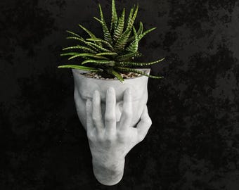 May include: A gray concrete planter shaped like a human face with a hand covering the face. The planter holds a green succulent plant. The background is a dark, textured surface.