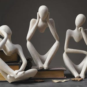 Pode incluir: Três esculturas abstratas de figuras sentadas em tons neutros. Cada figura está em uma pose diferente, com as mãos perto da cabeça ou do queixo, sugerindo contemplação. As esculturas estão sobre livros, criando uma exibição minimalista e artística.