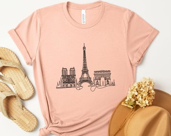 Puede incluir: Una camiseta rosa con un dibujo en línea negra de la Torre Eiffel, el Arco del Triunfo y la Catedral de Notre Dame. La camiseta está colocada sobre una superficie blanca con un sombrero de paja y sandalias.