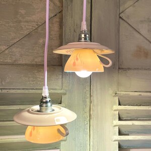 May include: Two white porcelain teacups with floral patterns are used as light fixtures, hanging from a light pink cord with white polka dots. The teacups are illuminated with light bulbs, casting a warm glow.