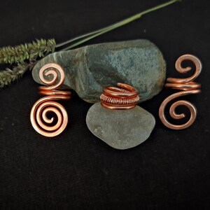 Puede incluir: Tres espirales de alambre de cobre para el cabello, cada una con un diseño de espiral diferente. Las espirales están dispuestas sobre una roca gris y una roca blanca.