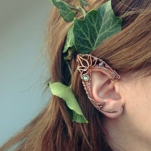 May include: A copper wire ear cuff with a floral design and green and purple gemstones. The cuff is worn on a person's ear.