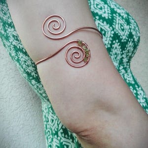 May include: Copper arm cuff featuring two spiral designs connected by a thin band. The lower spiral is adorned with small, light green beads. The jewelry is worn on the arm, against a background of a green and white patterned dress.