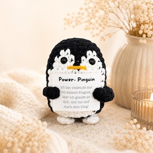 Könnte beinhalten: Ein handgefertigter Pinguin aus Häkelarbeit in Schwarz, Weiß und Gelb, der ein Schild hält. Auf dem Schild steht "Power- Pinguin" und enthält deutschen Text. Der Pinguin steht auf einer cremefarbenen Oberfläche, mit einer Kerze und einer Vase im Hintergrund.