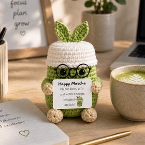 Könnte beinhalten: Eine gehäkelte grüne und weiße Matcha-Figur mit Brille, die ein Schild mit der Aufschrift "Happy Matcha" hält. Eine Tasse Matcha Latte und ein Notizbuch mit einer To-Do-Liste sind ebenfalls zu sehen.