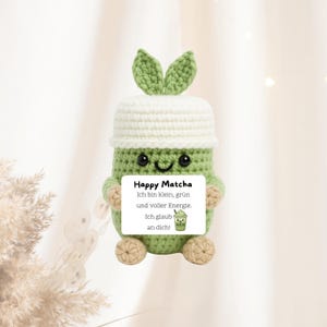 Könnte beinhalten: Ein gehäkelter grüner Matcha-Getränke-Charakter mit weißem Hut und grünen Blattakzenten. Die Figur hält ein Schild mit der Aufschrift "Happy Matcha" auf Deutsch, mit zusätzlichem Text und einer Matcha-Getränke-Illustration.