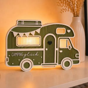May include: A decorative olive green and white wooden camper van lamp featuring a corrugated texture, a roof rack, a small heart on the door, and the text "CAMPING gl&uuml;ck" printed on the side. The lamp emits a warm glow from the windows.