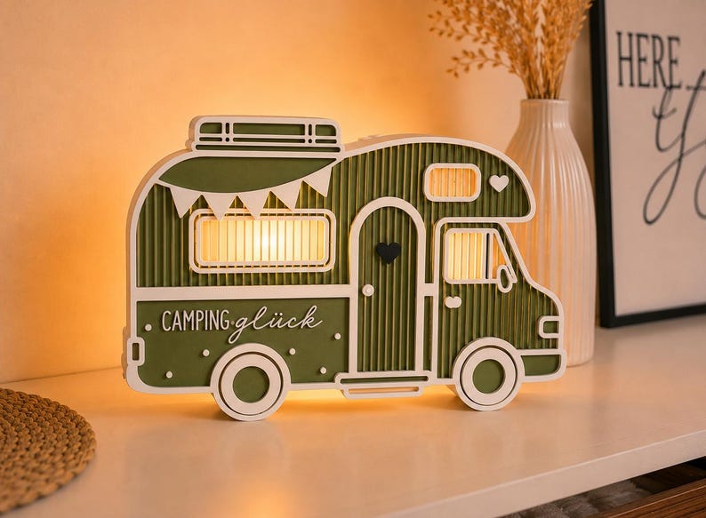 May include: A decorative olive green and white wooden camper van lamp featuring a corrugated texture, a roof rack, a small heart on the door, and the text "CAMPING gl&uuml;ck" printed on the side. The lamp emits a warm glow from the windows.