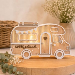 May include: A decorative wooden camper van light featuring a white frame and natural wood slats. The side of the camper displays the text "Camping gl&uuml;ck" in black script. A small dark heart is positioned on the door, and the interior is illuminated.