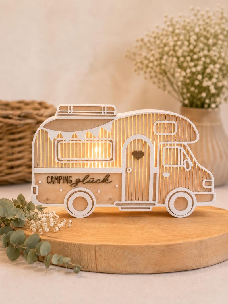 May include: A decorative wooden camper van light featuring a white frame and natural wood slats. The side of the camper displays the text "Camping gl&uuml;ck" in black script. A small dark heart is positioned on the door, and the interior is illuminated.