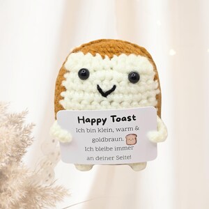 Peut inclure: Un personnage de toast crocheté avec un visage souriant, des yeux noirs et une palette de couleurs marron et blanche. Il tient une carte blanche avec le texte "Happy Toast" et du texte allemand. Le toast a de petits bras.