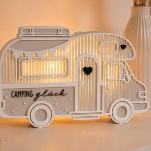 May include: A decorative light-up camper van ornament in white and light gray. The camper features a cutout design with vertical slats, a small black heart on the door, and the text "CAMPING gl&uuml;ck" printed on the side in black lettering.