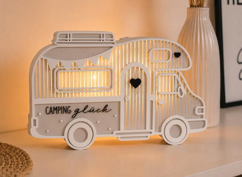 May include: A decorative light-up camper van ornament in white and light gray. The camper features a cutout design with vertical slats, a small black heart on the door, and the text "CAMPING gl&uuml;ck" printed on the side in black lettering.