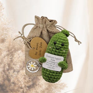 May include: A handmade green crochet pickle with a smiling face and a small sign that reads "Emotional Support Pickle." It is attached to a keychain with a daisy charm and a burlap gift bag with a heart-shaped tag.