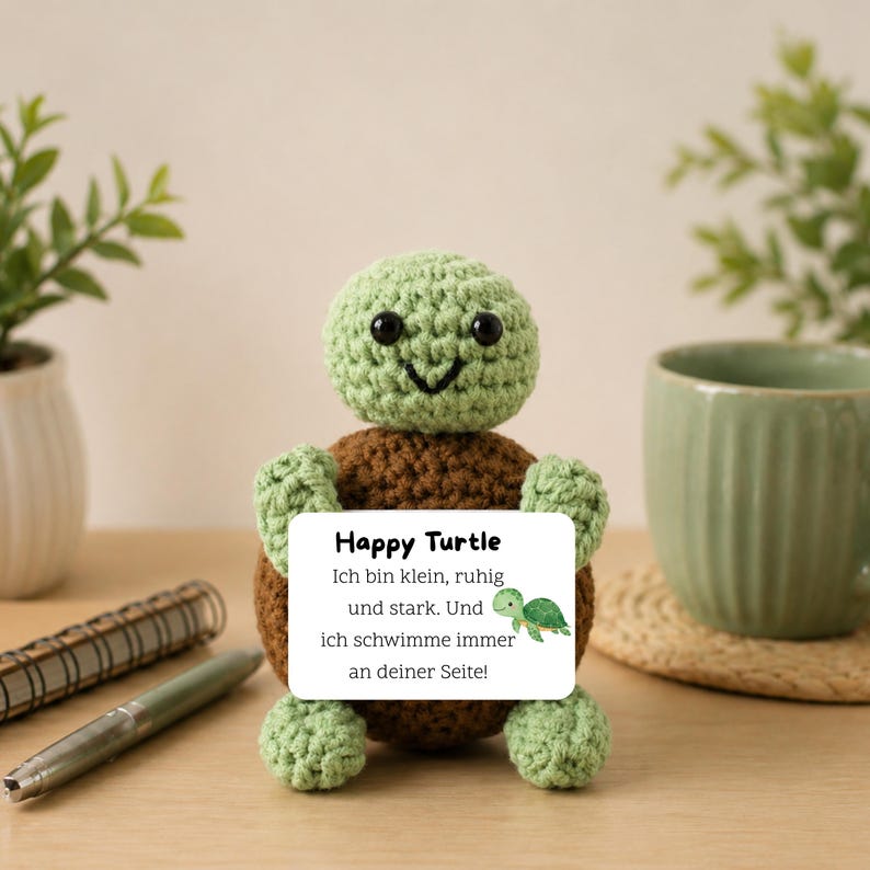 Eine handgefertigte Häkelschildkröte in Grün und Braun. Die Schildkröte hält ein weißes Schild mit dem Text "Happy Turtle" und deutschem Text. Eine grüne Tasse und ein Notizbuch befinden sich im Hintergrund.