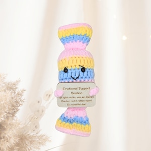 Peut inclure: Une figurine de bonbon crochetée à la main, aux couleurs vives. Le bonbon souriant porte une pancarte en bois avec l'inscription "Emotional Support Bonbon" en allemand. Rose, jaune et bleu.