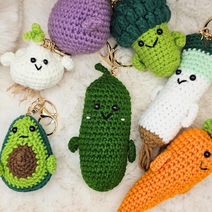 Puede incluir: Una colección de llaveros de verduras hechos a mano con ganchillo, que incluyen un aguacate verde, una cebolla morada, un pepinillo verde, un brócoli verde, un rábano blanco y una zanahoria naranja. Cada llavero tiene un cierre dorado y una cara sonriente.