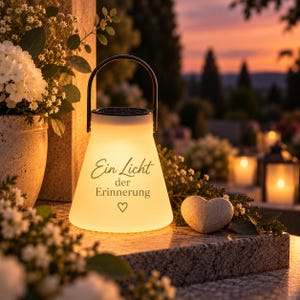 May include: A glowing, conical solar lantern with a black handle, featuring the German text 'Ein Licht der Erinnerung' and a small heart symbol printed on the front. The lantern emits a warm, soft light, illuminating the surrounding area.