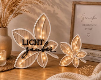 May include: Two leaf-shaped light-up decorations with white frames and light brown vertical-slatted centers. The larger leaf features the black text "LICHT zauber" across the front. Both pieces are illuminated by small, warm-toned LED lights.
