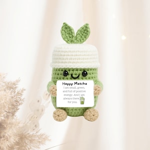 May include: A handmade crochet matcha drink plush toy, featuring a white lid and green leaves. The plushie has a smiling face and holds a sign that reads "Happy Matcha". The toy is green, beige, and white.