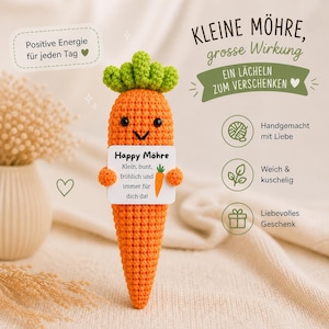 Könnte beinhalten: Eine handgefertigte orangefarbene Häkelkarotte mit grünem Oberteil. Die Karotte hat ein lächelndes Gesicht und hält ein Schild mit der Aufschrift "Happy Möhre". Das Schild hat auch eine kleine Karottenillustration. Der Text "Positive Energie für jeden Tag" befindet sich oben links.