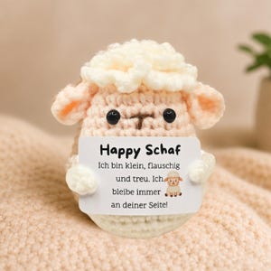Könnte beinhalten: Gehäkeltes Schaf in Creme- und Pfirsichtönen, hält eine weiße Karte mit deutschem Text. Das Schaf hat schwarze Knopfaugen und eine braune "T"-förmige Nase. Der Hintergrund ist ein weicher, strukturierter Stoff. Auf der Karte steht "Happy Schaf".