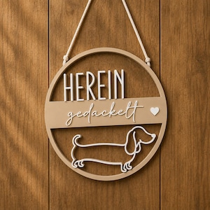 Könnte beinhalten: Ein rundes Wandschild aus Holz mit dem weißen Schriftzug HEREIN gedackelt und einer weißen Strichzeichnung eines Dackels. Das Schild hat eine hellbraune Oberfläche und ist an einer weißen Kordel befestigt, die oben am runden Rahmen sitzt.
