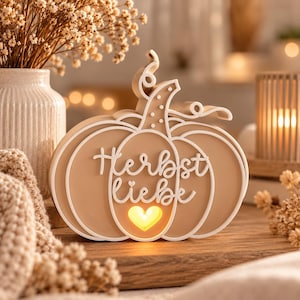 Könnte beinhalten: Eine beige, kürbisförmige Dekoleuchte mit weißen Umrandungen und einem geschwungenen Stiel. Der Schriftzug "Herbst liebe" ist in weißer Schreibschrift mittig angebracht, ergänzt durch eine herzförmige Aussparung mit warmem gelbem Licht.