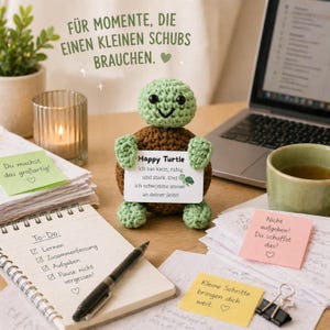 May include: A crocheted turtle figurine with a sign that reads "Happy Turtle." The turtle is brown and green, with a smiling face. The image includes a laptop, a cup of tea, and sticky notes with motivational messages.