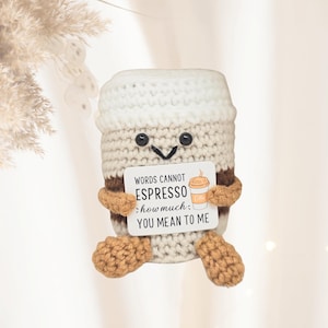 Puede incluir: Una taza de café de ganchillo con la parte superior blanca, el centro marrón y la parte inferior beige. La taza tiene una cara sonriente y sostiene un letrero que dice "WORDS CANNOT ESPRESSO how much YOU MEAN TO ME."
