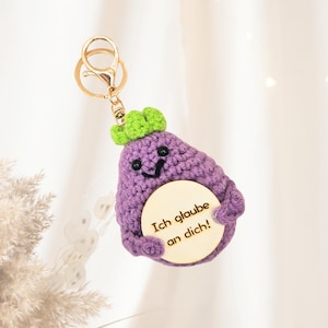 Puede incluir: Un llavero de berenjena de ganchillo morado hecho a mano con una parte superior verde y un disco de madera que dice "Ich glaube an dich!" en texto negro. El llavero tiene un cierre dorado.