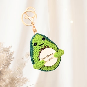 Puede incluir: Un llavero de aguacate tejido a mano, verde y marrón, con un cierre dorado. Sostiene un disco de madera con el texto alemán "Ich glaube an dich!". Un accesorio encantador y original.